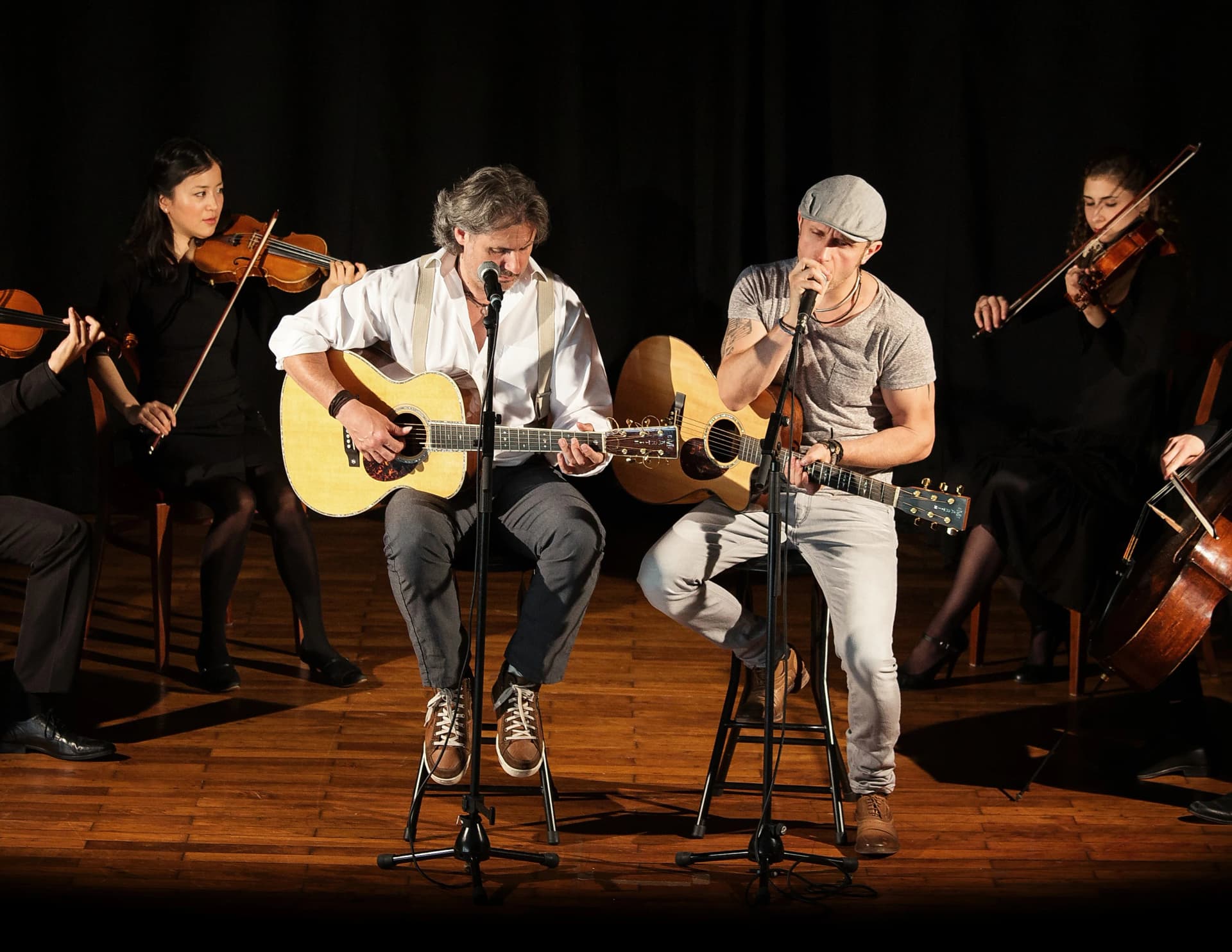 Zwei Musiker mit Akustikgitarren sitzen auf Barhocker an Mikrofonen und singen, umrahmt von einem Streichquartett mit Geigen und Cello auf einer Bühne.
