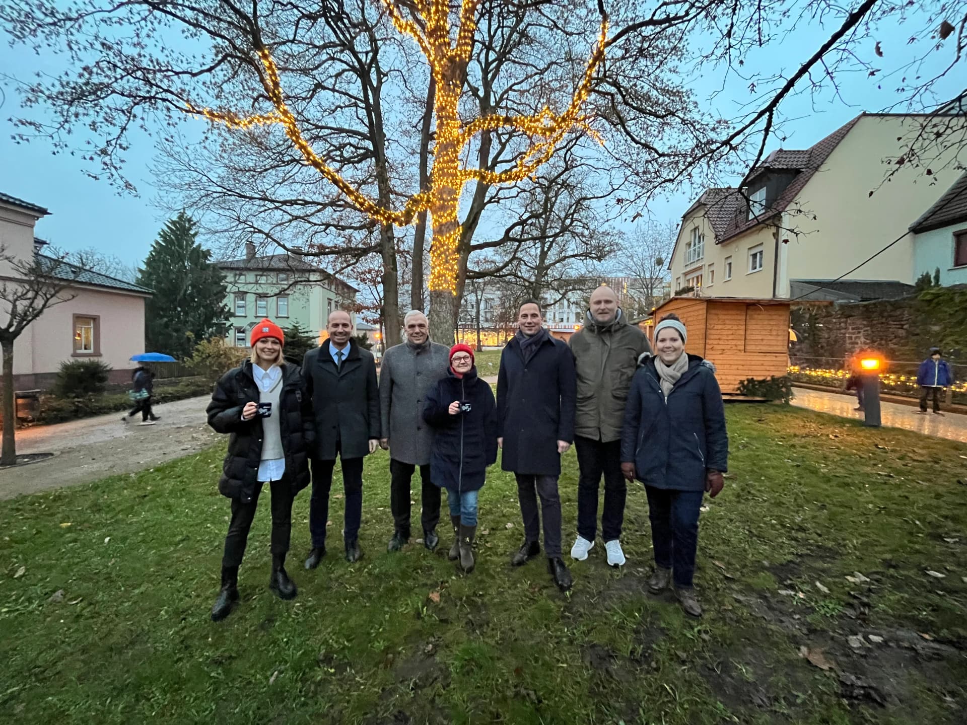 Gruppe von sieben Personen steht abends vor einem mit Lichterketten beleuchteten Baum in einer Grünanlage