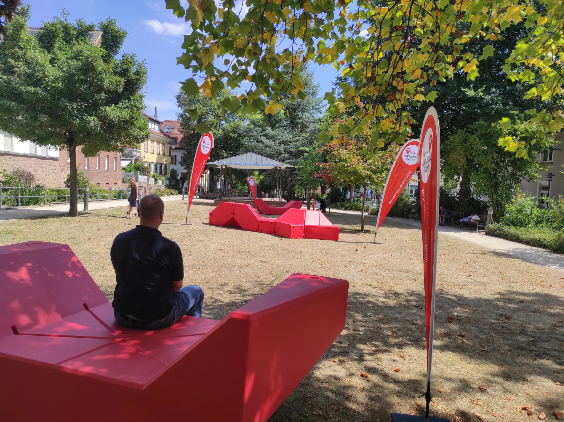 Park mit roten Sitzmöbeln und Werbefahnen 'Lohrer Lieblingsplatz', ein Mann sitzt auf einer Bank im Schatten der Bäume.