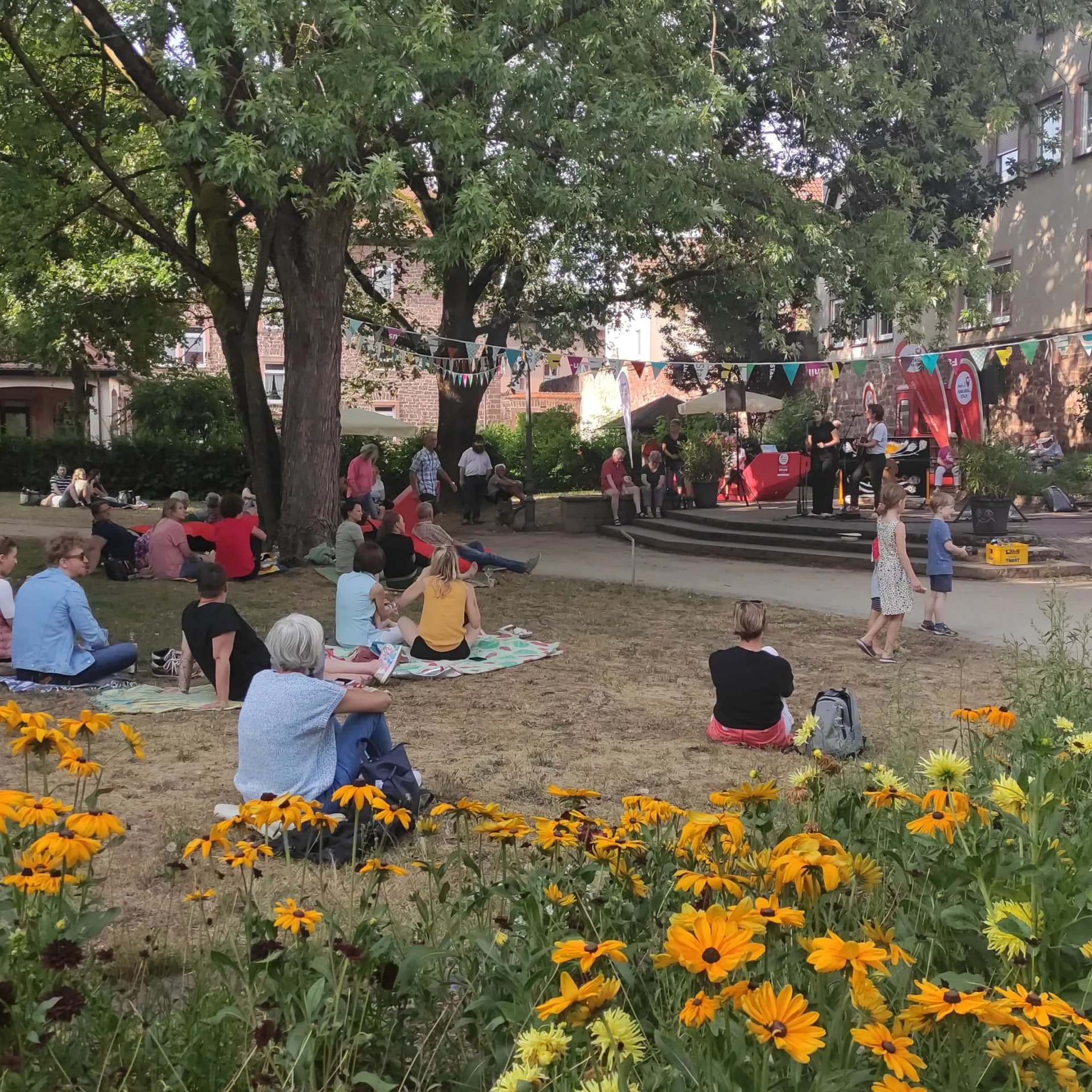 Menschen sitzen auf Decken im Grünen unter Bäumen und hören einem Konzert auf einer kleinen Bühne zu, im Vordergrund gelbe Blumen