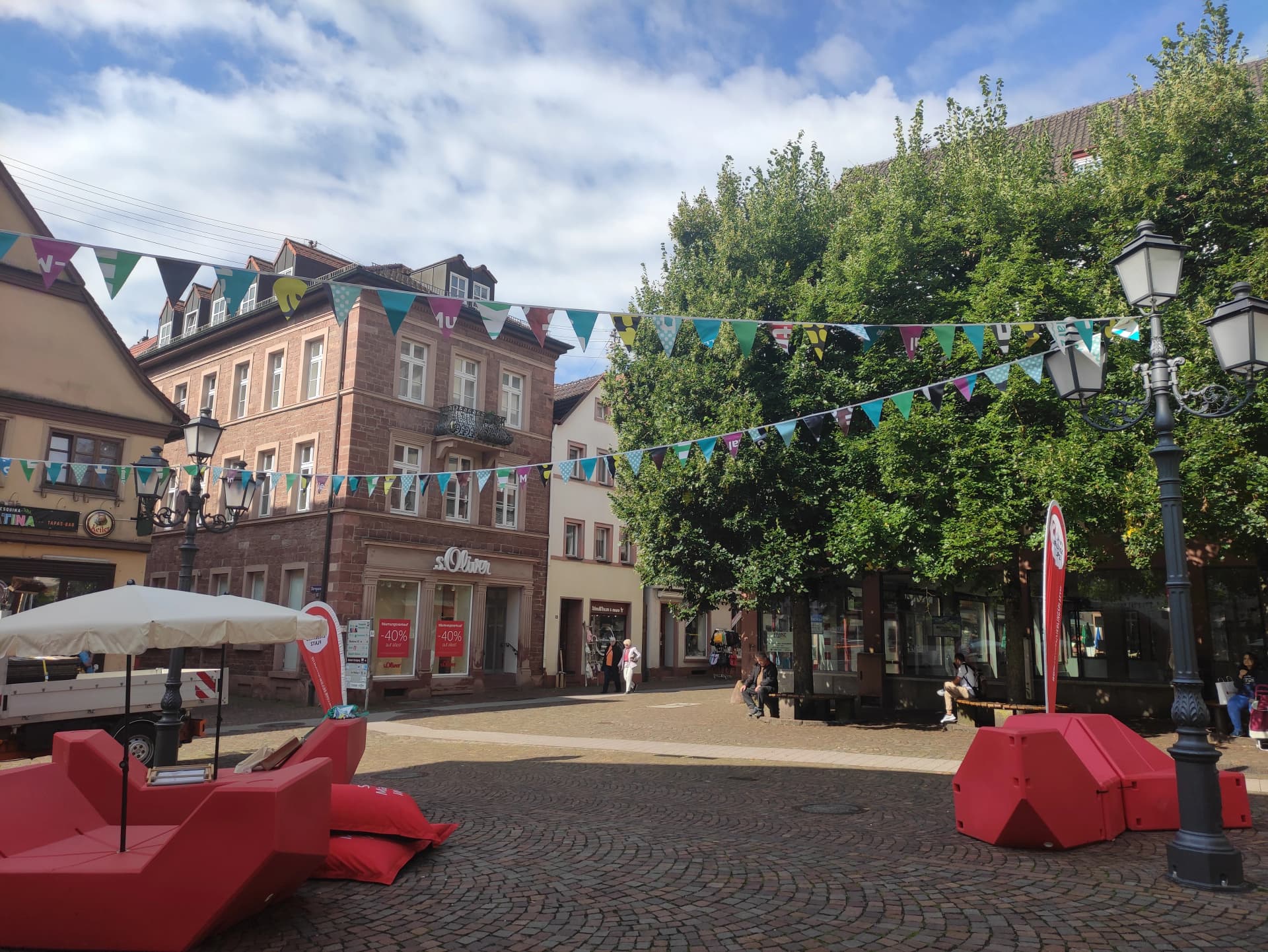 Fußgängerzone mit historischen Fachwerk- und Sandsteinhäusern, Wimpelgirlanden über der Straße und roten Sitzmöbeln im Vordergrund