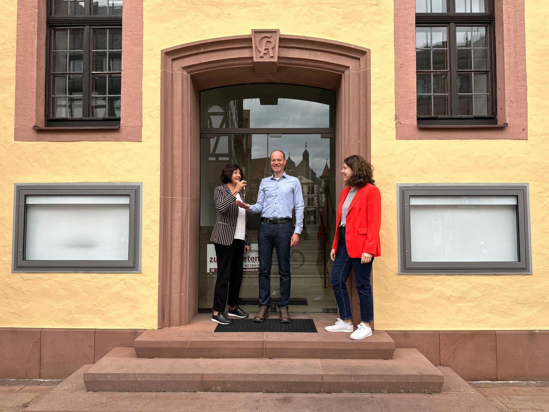 Zwei Frauen und ein Mann stehen lächelnd vor einem historischen Hauseingang mit Sandsteinportal, im Vordergrund eine symbolische Schlüsselübergabe.