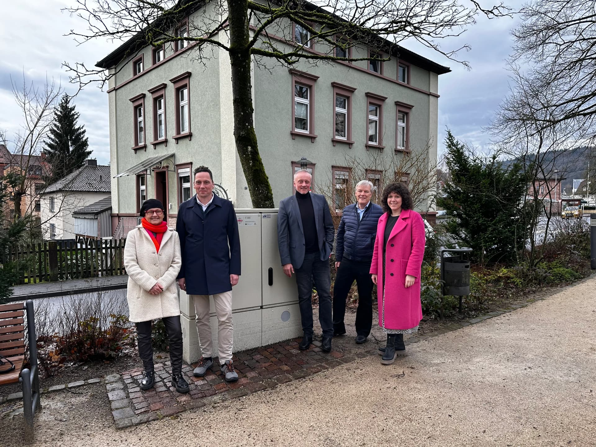 Fünf Personen stehen vor einem grün verputzten Mehrfamilienhaus neben einem Stromverteilerkasten im Freien.