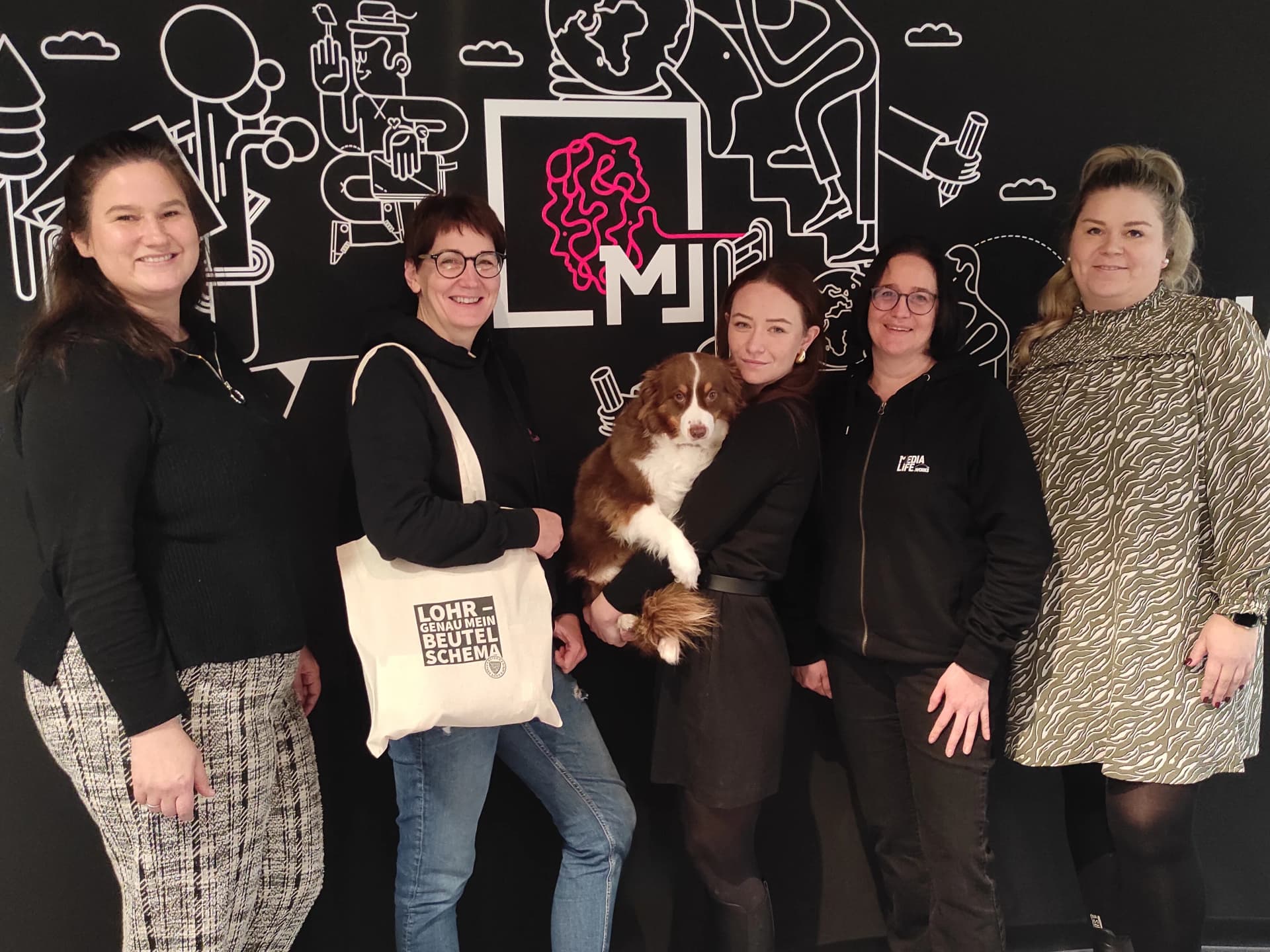 Fünf Frauen und ein Hund stehen vor einer bemalten Wand mit Media-Life-Logo, eine hält eine Stofftasche mit der Aufschrift 'Lohr – genau mein Beutelschema'