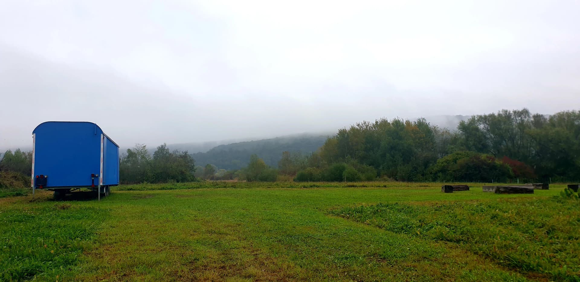 Blauer Bauwagen auf einer Wiese vor bewaldeten, nebligen Hügeln