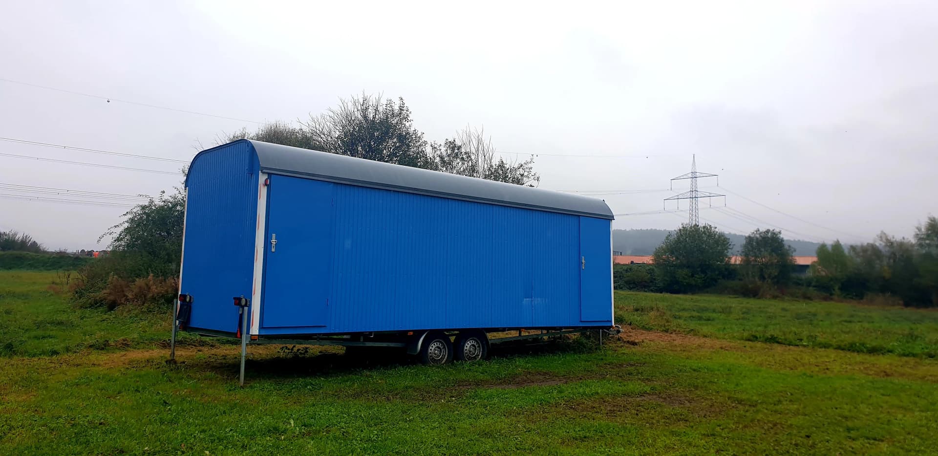 Blauer Bauwagen auf einer Wiese mit Bäumen und Strommast im Hintergrund
