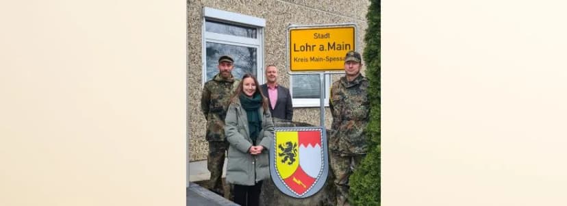 Vier Personen, zwei in Bundeswehruniform, stehen vor dem Ortsschild Stadt Lohr a. Main, Kreis Main-Spessart, mit Stadtwappen.