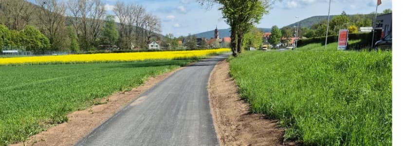 Asphaltierter Radweg zwischen Feldern mit blühendem Raps, im Hintergrund ein Ort mit Kirchturm vor Hügeln