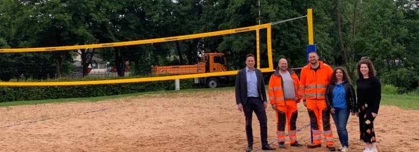 Fünf Personen, darunter zwei in orangener Arbeitskleidung, stehen auf einem Beachvolleyballplatz mit Sandfeld und Netz