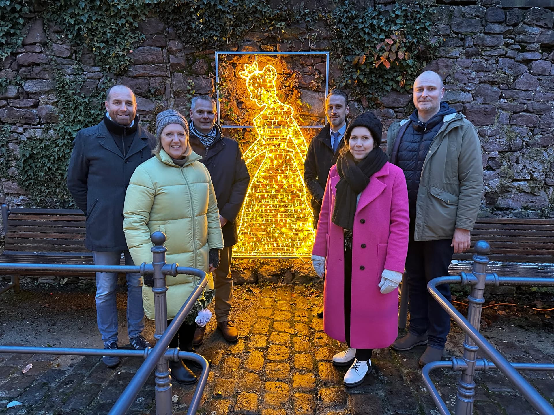 Sechs Personen stehen vor einer beleuchteten Schneewittchen-Figur aus Lichterketten an einer efeubewachsenen Steinmauer.