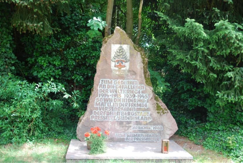 Gedenkstein mit Inschrift zum Andenken an die Gefallenen der Weltkriege und Verstorbenen der Bergstadt Pressnitz, umgeben von Bäumen, mit Blumen und Grablicht d