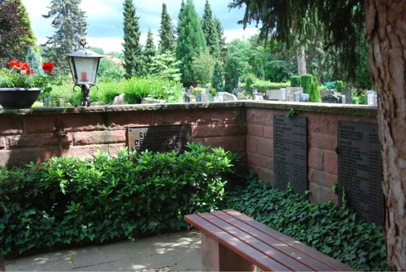 Gedenkstätte auf einem Friedhof mit Gedenktafeln an einer Mauer, umgeben von Sträuchern und einer Bank