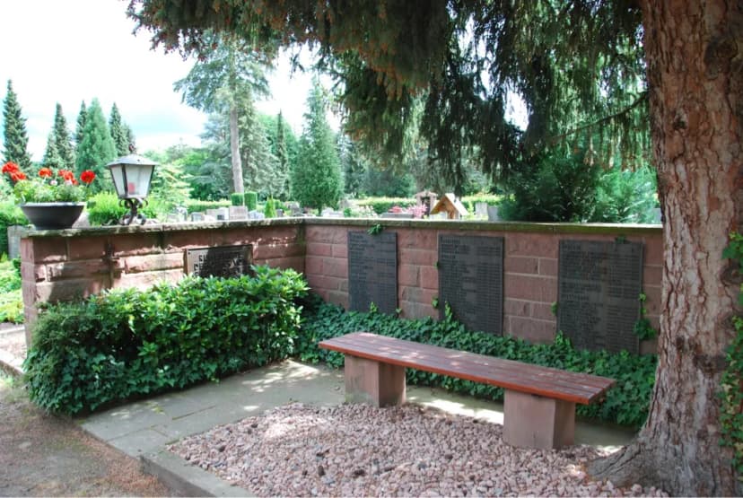 Gedenkstätte auf einem Friedhof mit Namenstafeln an einer Sandsteinmauer, davor eine Sitzbank und Sträucher unter einem großen Baum.