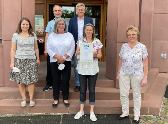 Sechs Personen stehen für ein Gruppenfoto vor dem Eingang eines Gebäudes, eine Frau hält eine Urkunde in der Hand.