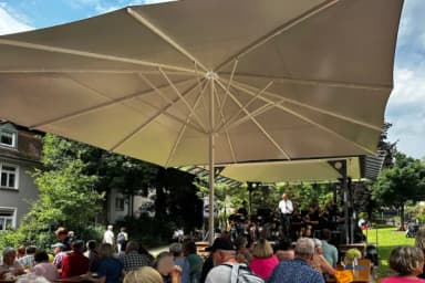 Besucher sitzen unter großen Sonnenschirmen bei einem Konzert im Freien