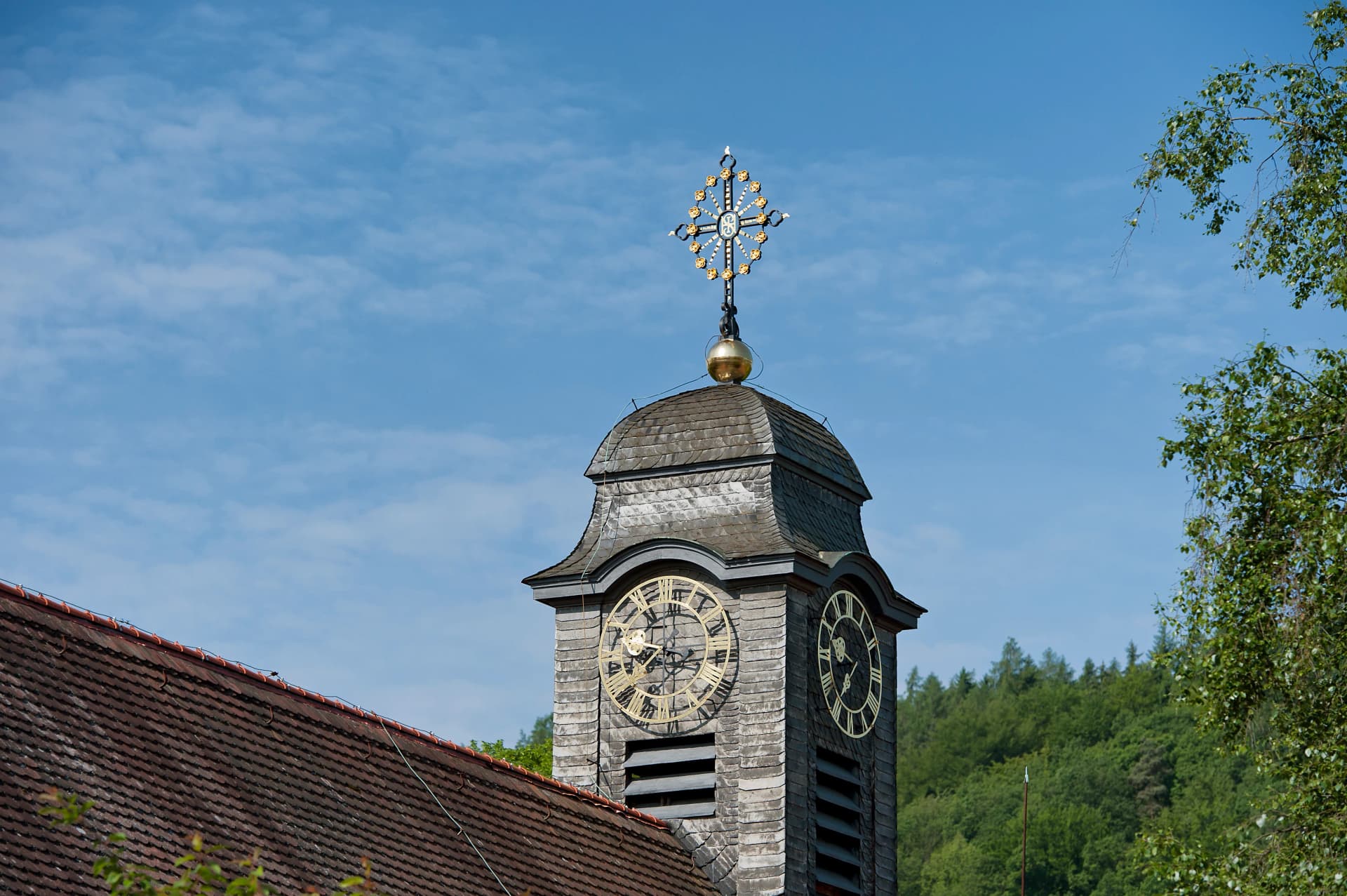 Kirchturm mit schiefergedecktem Dach und zwei Zifferblättern einer Turmuhr, bekrönt von einer goldverzierten Kreuzspitze auf goldener Kugel, vor blauem Himmel und Bäumen.