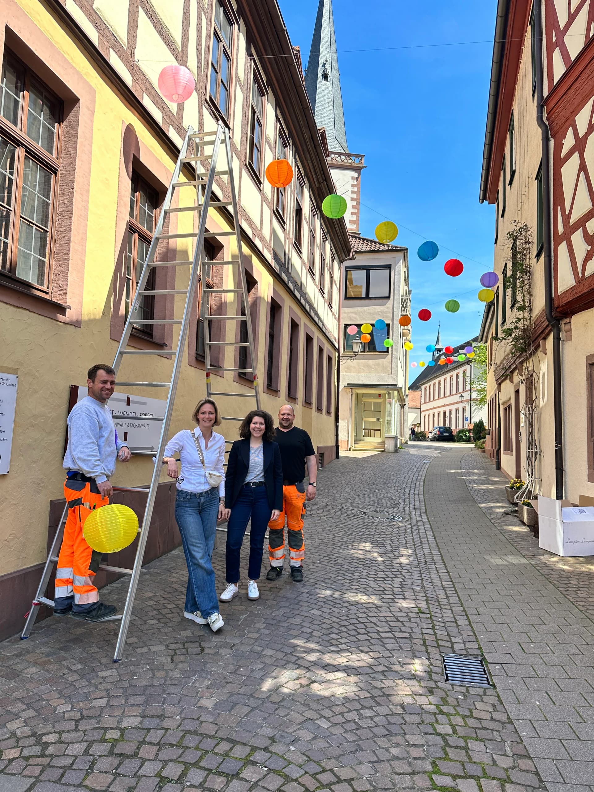 Gruppe von Personen, teils in orangefarbener Warnkleidung, steht neben einer Rampe in einer historischen Fachwerkstraße, darüber bunte Lampions gespannt.