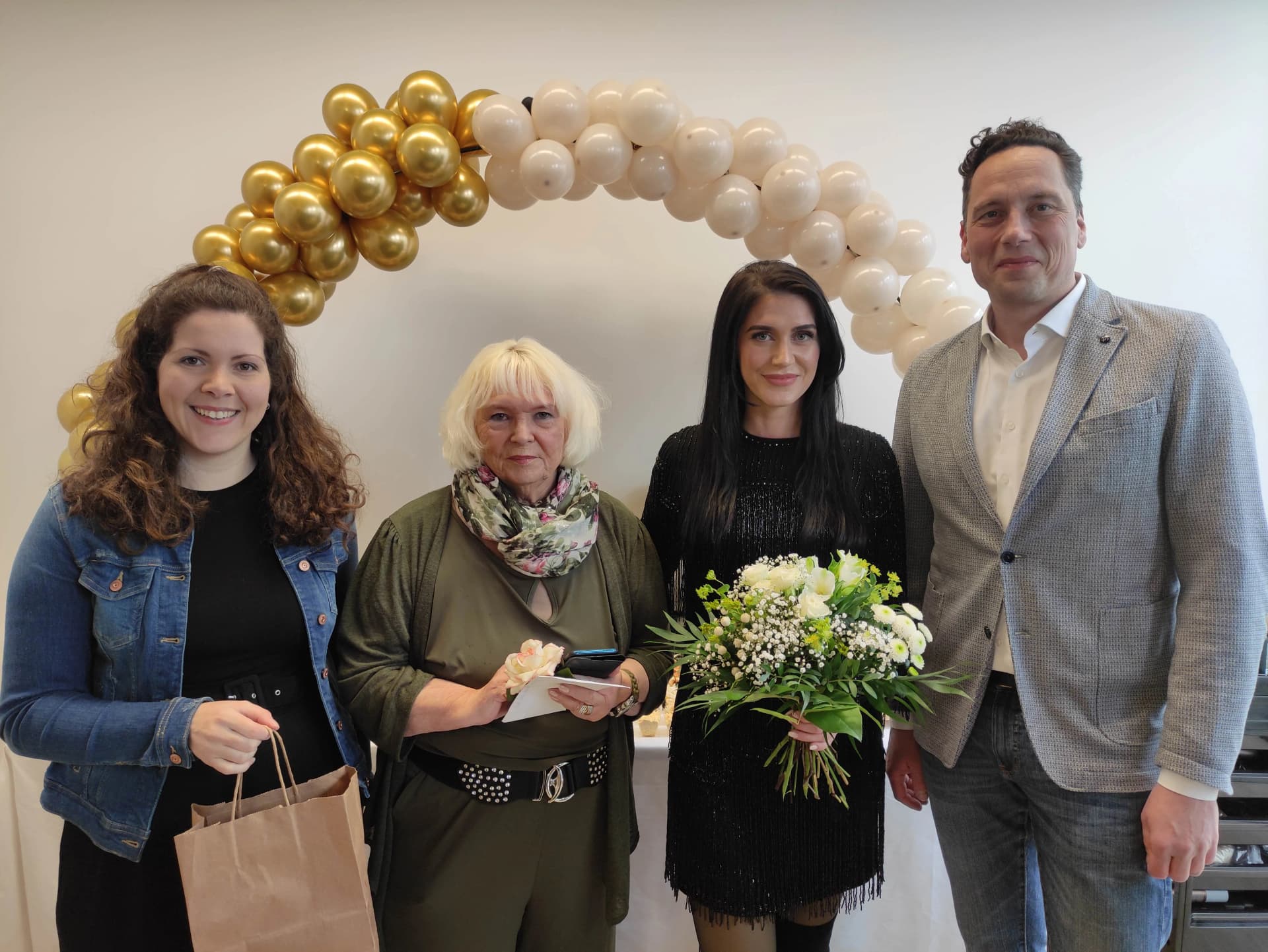 Vier Personen stehen lächelnd unter einem Ballonbogen aus goldenen und weißen Ballons, eine Frau hält einen Blumenstrauß.