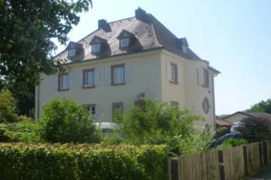 Historisches Gebäude mit Mansarddach, hellem Putz und grünen Fensterläden, umgeben von Hecken und Bäumen