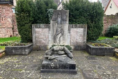 Steinernes Kriegerdenkmal mit Reliefdarstellung einer Figur und liegender Skulptur eines gefallenen Soldaten, umgeben von Hecken.