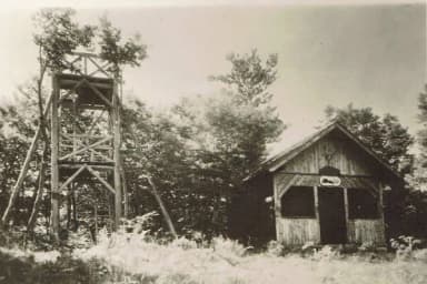 Historische Schwarzweißaufnahme eines hölzernen Aussichtsturms neben einer Holzhütte im Wald