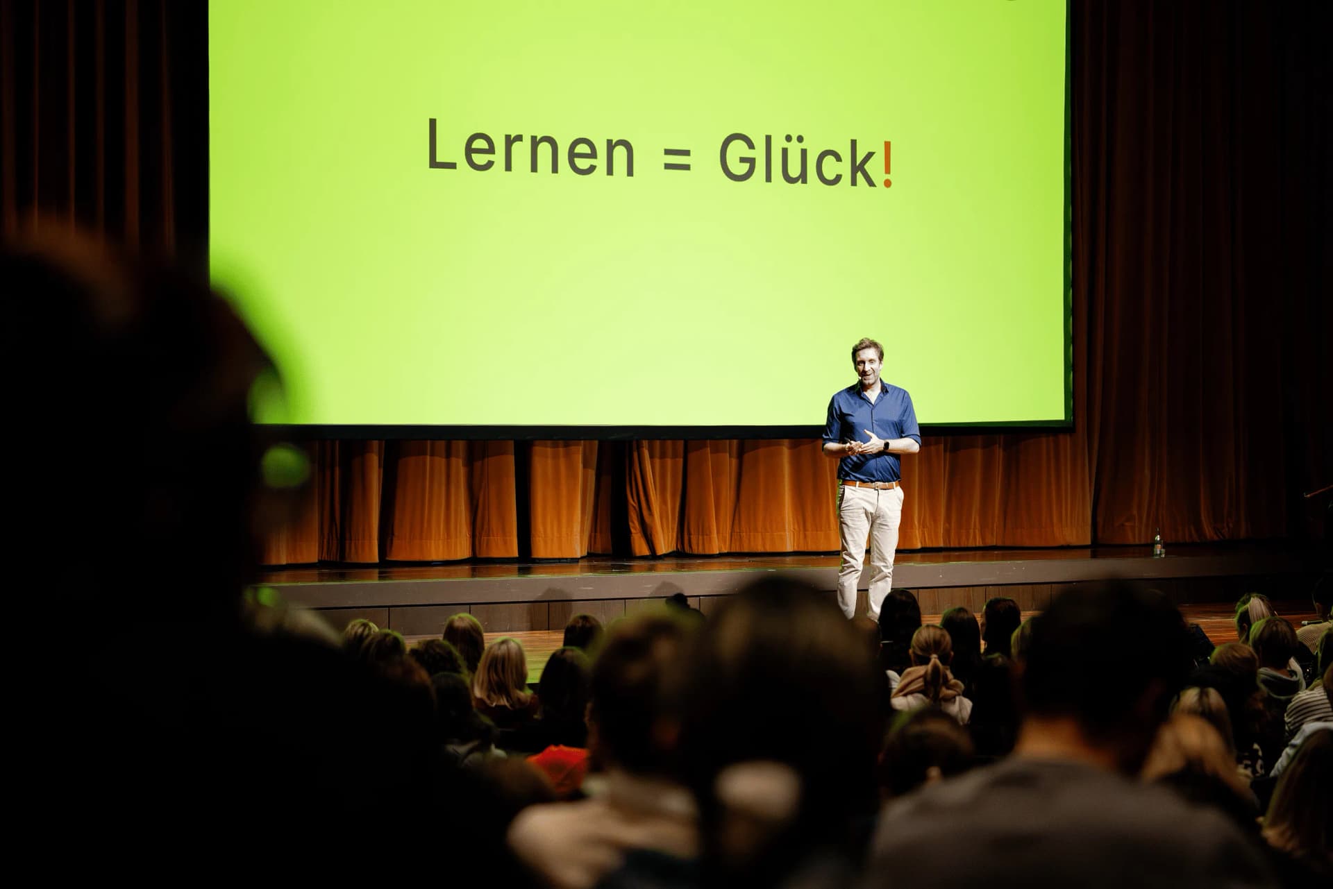 Ein Mann steht vor Publikum auf einer Bühne, im Hintergrund eine Leinwand mit der Aufschrift „Lernen = Glück!".