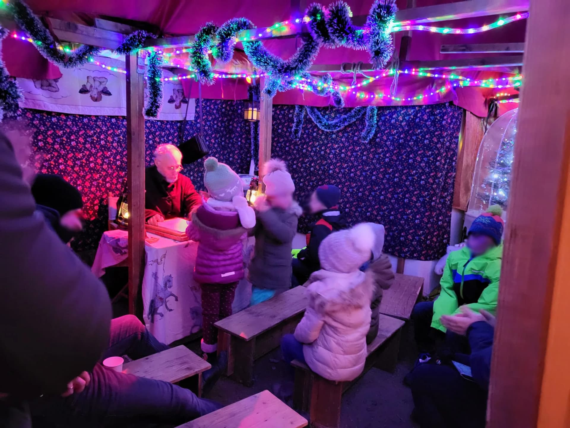 Kinder sitzen auf Bänken in einem festlich mit Lichterketten geschmückten Märchenzelt und lauschen einem Erzähler an einem Tisch.