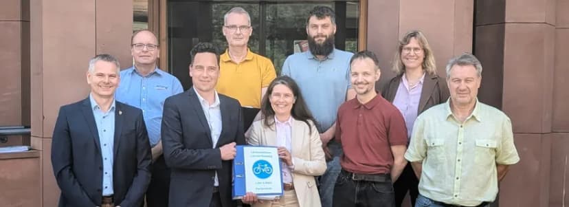 Gruppe von neun Personen vor einem Gebäude, in der Mitte hält eine Frau einen blauen Ordner mit der Aufschrift Lückenschluss Lohrtal-Radweg.