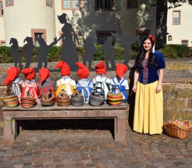 Das Lohrer Schneewittchen und ihre sieben Zwerge sitzen auf einer Bank direkt vor Schneewittchens Geburtshaus, dem Lohrer Schloss, wo sich heute das Spessartmuseum befindet.