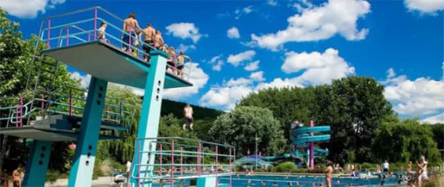 Sprungturm im Freibad mit Badegästen, im Hintergrund Wasserrutsche und Schwimmbecken vor bewaldeten Hügeln