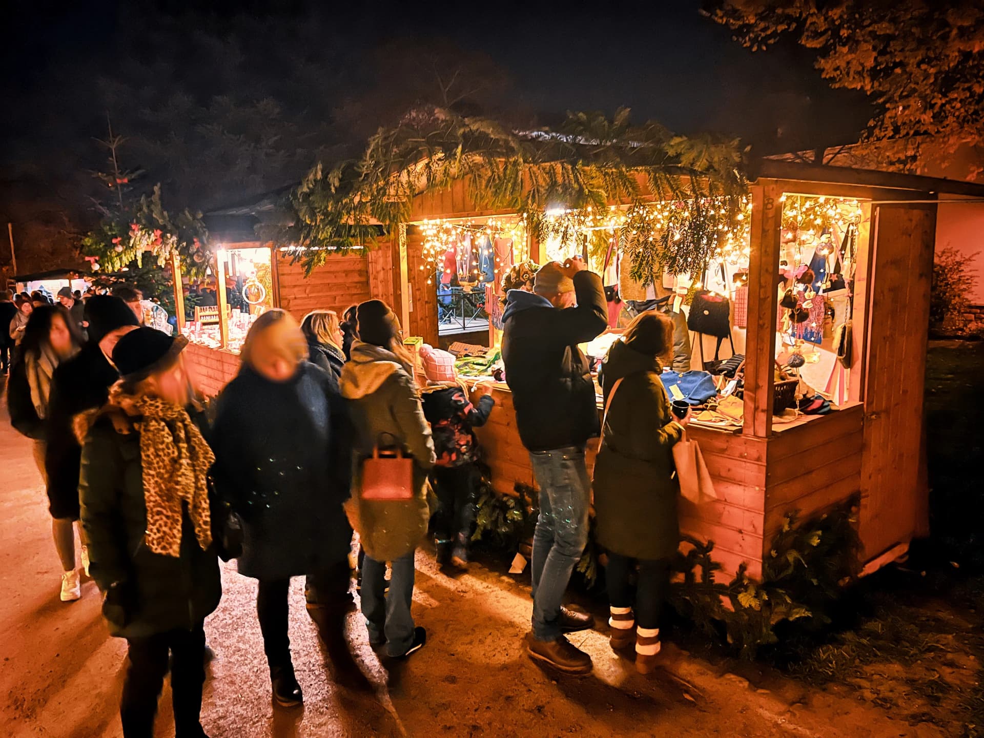 Besucher stehen abends an einem beleuchteten Weihnachtsmarktstand mit Textilwaren.