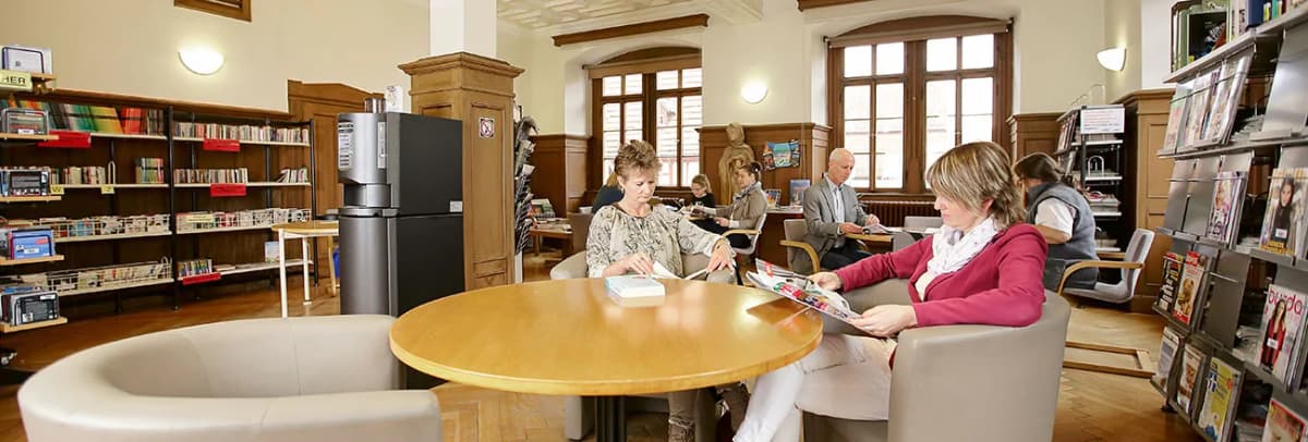Leseraum einer Bibliothek mit Regalen, Zeitschriften und Besuchern an Tischen