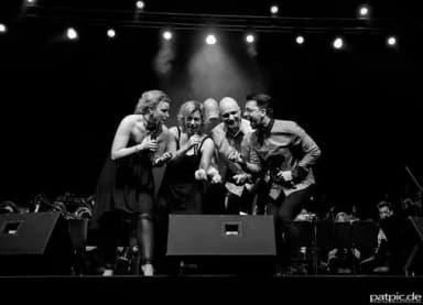 Vier Sängerinnen und Sänger mit Mikrofonen stehen eng beieinander auf einer Bühne und singen, im Hintergrund ein Orchester.