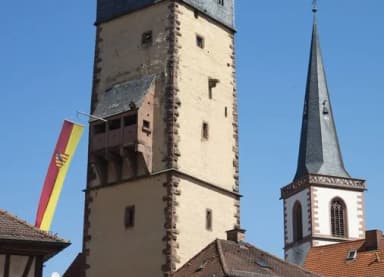 Historischer Wehrturm mit Fahne und benachbarter Kirchturmspitze vor blauem Himmel
