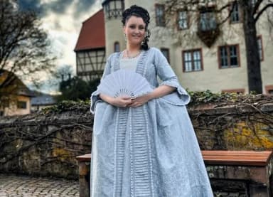 Frau in historischem hellblauem Barockkleid mit Fächer vor einem Fachwerkgebäude und einer Schlossmauer.