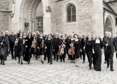 Orchestermitglieder in schwarzer Kleidung mit Instrumenten gehen vor einer Kirche über einen gepflasterten Platz.