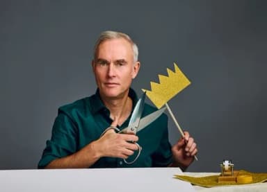 Mann sitzt an einem Tisch, hält eine Schere und eine goldene Papierkrone am Stiel in den Händen.