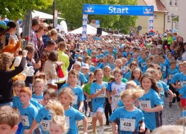 Kinder mit Startnummern laufen unter einem Startbogen bei einem Volkslauf, Zuschauer stehen am Straßenrand.