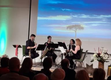 Vier Musiker spielen Saxophon auf einer Bühne vor einer Leinwand mit Naturmotiv, im Vordergrund sitzendes Publikum.