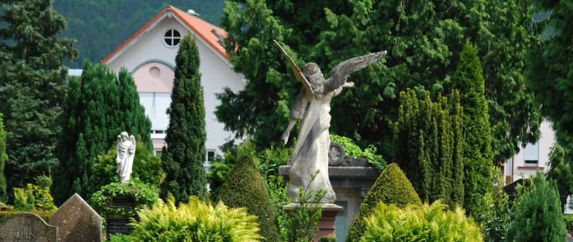 Engelsstatuen zwischen Grabsteinen und Bäumen auf einem Friedhof, im Hintergrund ein Gebäude mit rotem Dach