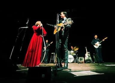 Band auf einer Bühne, eine Sängerin im roten Kleid und ein Sänger mit Gitarre am Mikrofon, im Hintergrund Schlagzeug und Gitarrist.