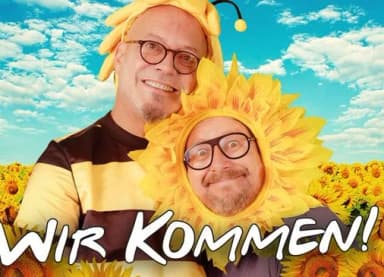 Zwei Männer verkleidet als Biene und Sonnenblume vor einem Sonnenblumenfeld, darunter der Schriftzug „Wir kommen!“