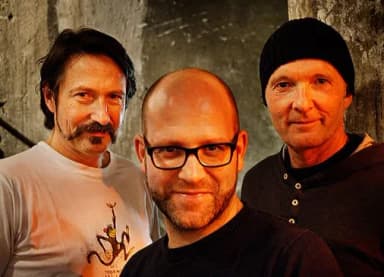 Drei Männer stehen nebeneinander vor einer Steinwand und blicken lächelnd in die Kamera.