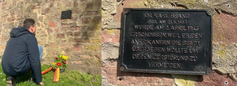Mann kniet mit Blumen vor einer Gedenktafel an einer Mauer; die Tafel erinnert an Dr. Karl Brand, der am 2. April 1945 erschossen wurde, weil er die Stadt den A