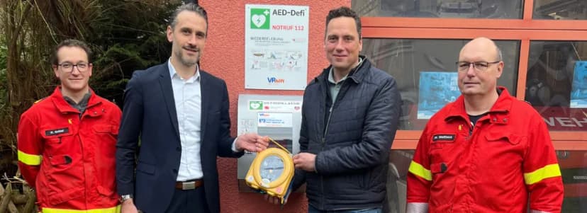Vier Personen, zwei in Feuerwehrkleidung, stehen vor einem an der Wand montierten Defibrillator-Kasten mit Aufschrift AED-Defi.