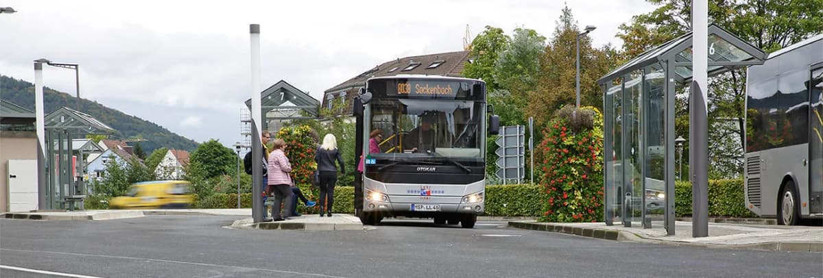 Bus an einer Bushaltestelle mit wartenden Fahrgästen, im Hintergrund Häuser und Berge