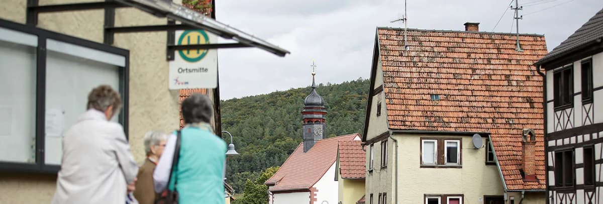 Ortsansicht mit Kirchturm und Fachwerkhäusern vor bewaldetem Hügel, im Vordergrund ein Schild „Ortsmitte“ und Passanten