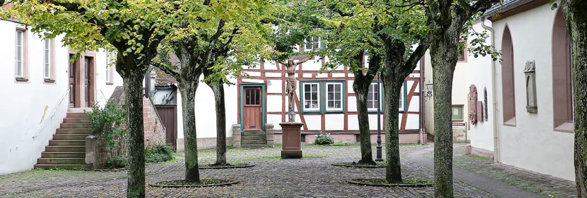 Von Bäumen umgebener Kirchplatz mit Fachwerkhaus und Kreuz-Statue auf Sockel