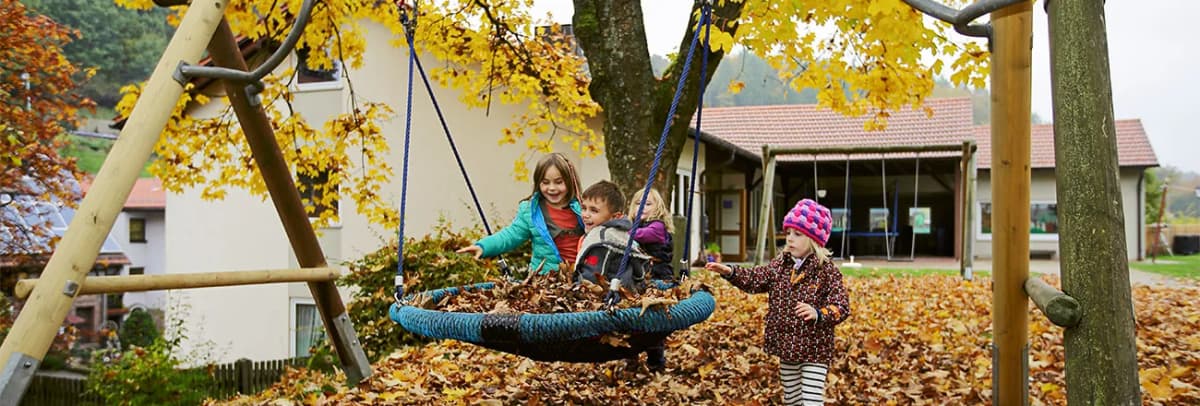 Kinder spielen auf einer Nestschaukel voller Herbstlaub auf einem Spielplatz vor Gebäuden