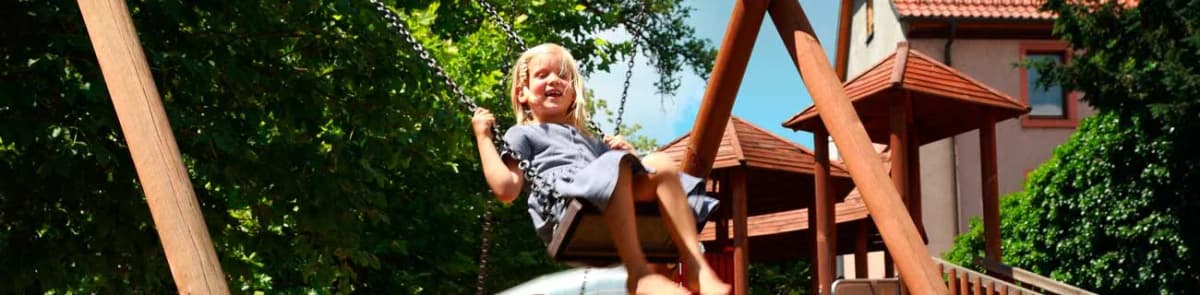 Kind schaukelt lachend auf einem Spielplatz mit Holzspielgeräten und Rutsche